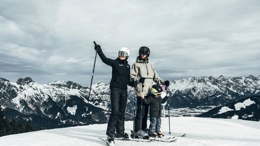 Eine Familie mit Kind steht auf der verschneiten Piste; die Frau weist in die Ferne.