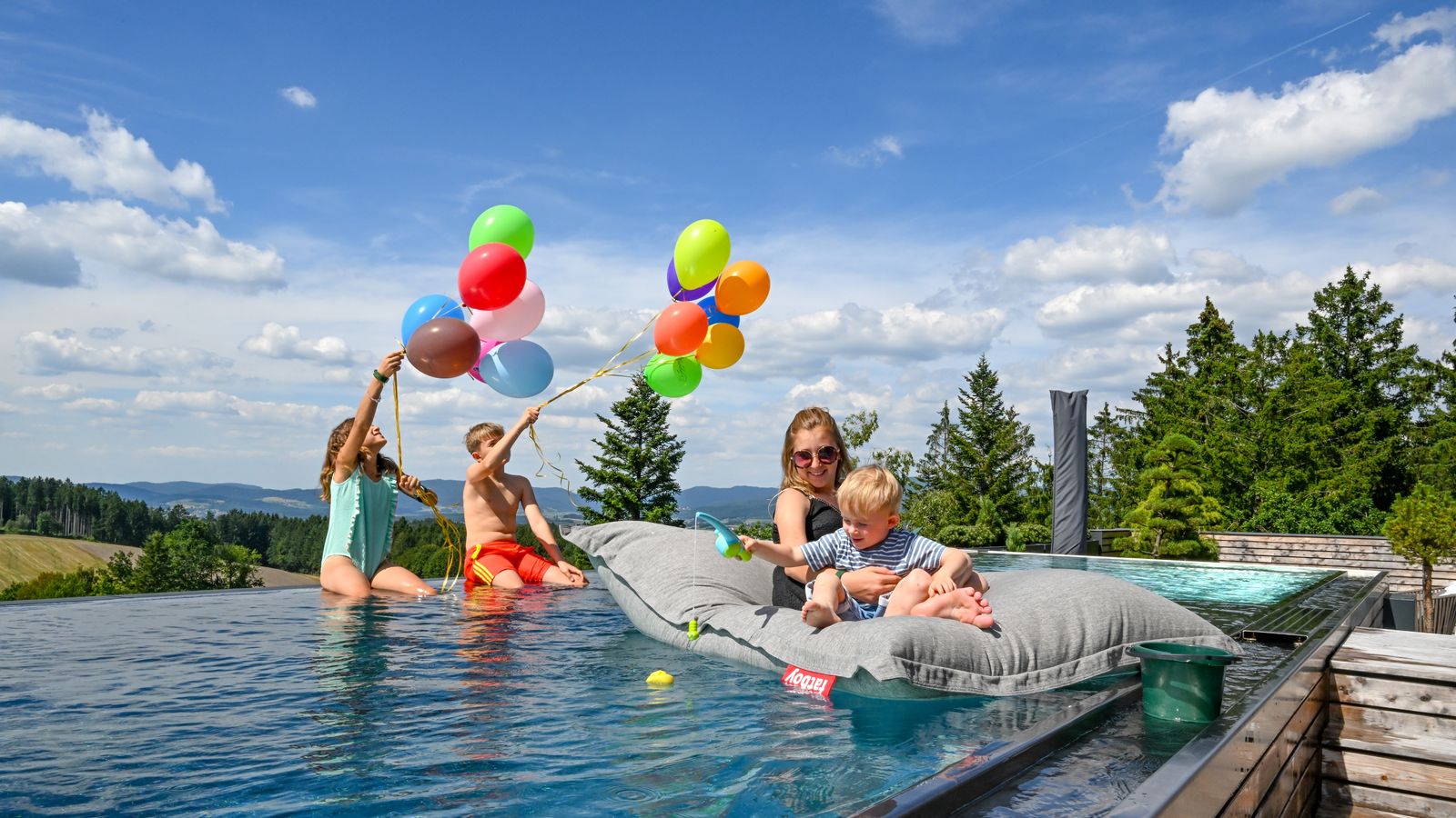 Kinder genießen den privaten Pool des Penthouse im ULRICHSHOF: Zwei Kinder spielen fröhlich mit bunten Luftballons im Wasser, während die große Schwester mit einem Kleinkind entspannt auf einer Luftmatratze treibt. Im Hintergrund erstreckt sich der Blick über Wälder und Hügel des Bayerischen Waldes.