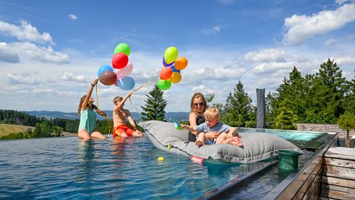 Kinder genießen den privaten Pool des Penthouse im ULRICHSHOF: Zwei Kinder spielen fröhlich mit bunten Luftballons im Wasser, während die große Schwester mit einem Kleinkind entspannt auf einer Luftmatratze treibt. Im Hintergrund erstreckt sich der Blick über Wälder und Hügel des Bayerischen Waldes.