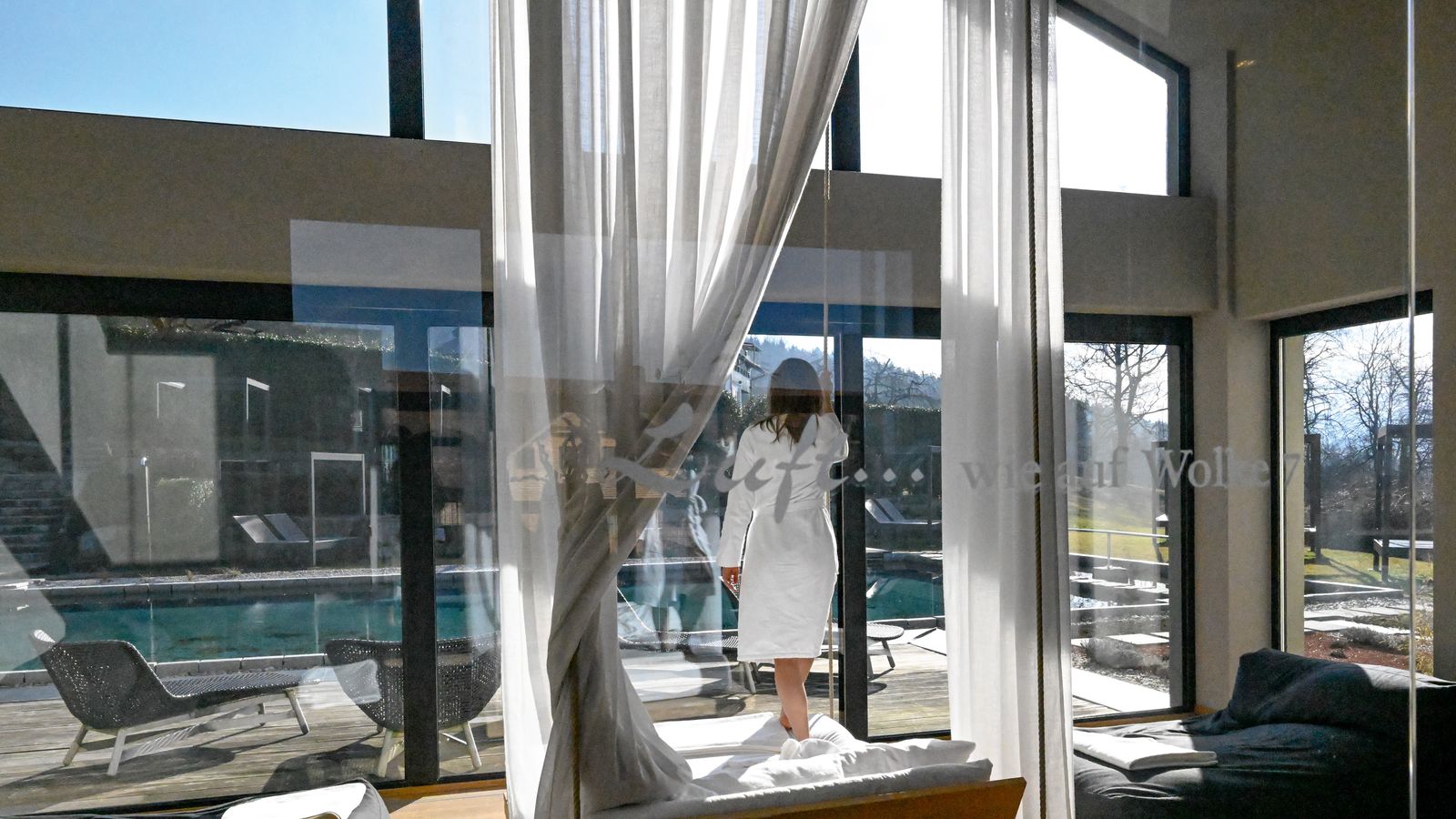 Frau im Bademantel steht im lichtdurchfluteten Eltern-SPA des ULRICHSHOF und blickt durch große Panoramafenster auf den Außenpool und die Natur. Vorhänge und gemütliche Liegen schaffen eine entspannte Wohlfühlatmosphäre.