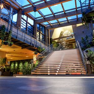 Großzügiges Lobby-Foyer des ULRICHSHOF mit breiter Treppe, Glasdach, hängenden Pflanzen und modernen Sitzbereichen. Die warme Beleuchtung betont die Kombination aus Holz, Stahl und Glas.