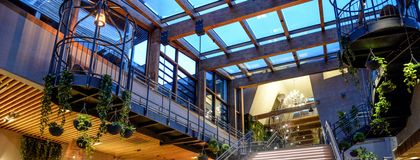Großzügiges Lobby-Foyer des ULRICHSHOF mit breiter Treppe, Glasdach, hängenden Pflanzen und modernen Sitzbereichen. Die warme Beleuchtung betont die Kombination aus Holz, Stahl und Glas.