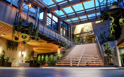 Großzügiges Lobby-Foyer des ULRICHSHOF mit breiter Treppe, Glasdach, hängenden Pflanzen und modernen Sitzbereichen. Die warme Beleuchtung betont die Kombination aus Holz, Stahl und Glas.
