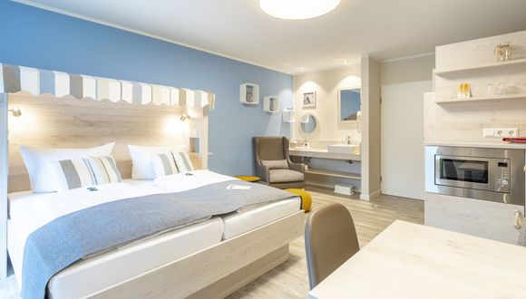 Links das Doppelbett mit Strandkorb-Stil, dahinter die Waschecke und rechts die Sitzecke und Schränke mit der Mikrowelle