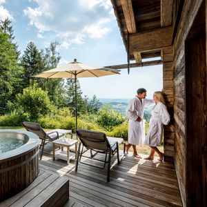 Pärchen steht im Luxus-Chalet auf der eigenen Sonnenterrasse neben Whirlpool und traumhaften Ausblick über den Bayerischen Wald.