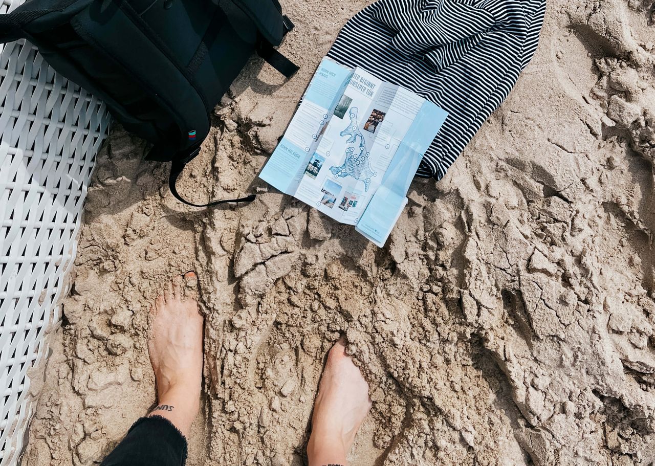 Nahaufnahme von zwei Füßen im Sand, daneben liegen ein Rucksack, ein Oberteil und ein Flyer für nachhaltigen Urlaub